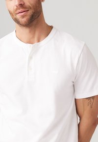 Witte katoenen henley shirt met korte mouwen, drie knopen en een ronde halslijn, met subtiele branding op de borst. Glad textuur.