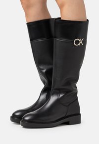 Stivali neri al ginocchio realizzati in pelle liscia. Presentano una punta arrotondata, un tacco a blocco e un logo "CK" in argento sul lato esterno.