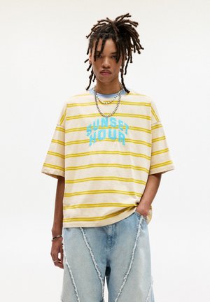 T-shirt beige a righe con accenti gialli e blu, con la scritta "SUNSET HOUR" ricamata in blu. Abbinato a pantaloni corti in denim strappati.