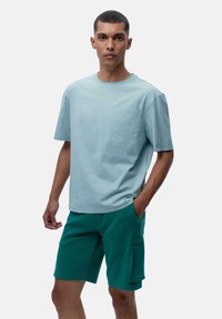 Šviesiai mėlynas medvilninis marškinėliai su apvalia iškirpte ir priekinėmis kišenėmis, derinami su žaliais šortais, turinčiais šoninę kišenę ir siuvinėjimo detales.