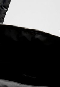 Crna najlon torba s glatkom teksturom, sadrži srebrni zatvarač duž gornjeg dijela. Unutrašnjost prikazuje logo i minimalno označavanje.