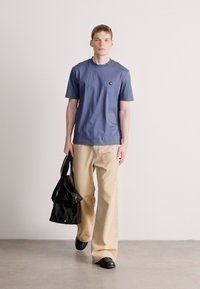 Μπλε βαμβακερή μπλούζα με στρογγυλή λαιμόκοψη και μικρό λογότυπο, συνδυασμένη με μπεζ παντελόνι φαρδιάς γραμμής και μαύρα παπούτσια, κρατώντας μαύρη τσάντα.