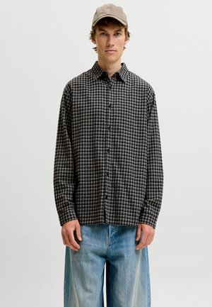 Jeune homme portant une casquette beige, une chemise à carreaux noirs et blancs boutonnée, et un jean bleu clair ample, debout devant un fond clair uni.