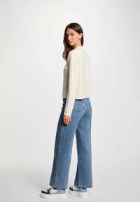 Pullover crema con colletto alto, abbinato a jeans a gamba larga di colore blu chiaro. Il modello indossa sneakers bianche con dettagli blu.