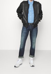 Svart läderbombarjacka, ljusblå skjorta, mörka jeans med uppvikt fåll, vita sneakers med grå detaljer, stående mot en neutral bakgrund.