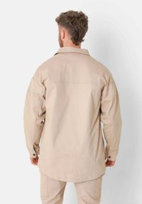 Camisa de manga larga beige con yugo trasero, hombros caídos y puños abotonados. La tela parece resistente y tiene una textura suave.