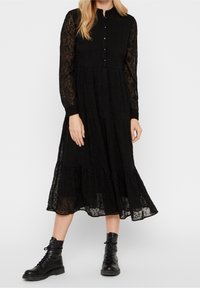 Kvinna klädd i en svart långärmad midiklänning med genomskinliga ärmar och svarta snörstövlar, stående mot en enfärgad bakgrund.