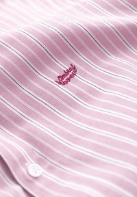 Tissu rose avec de fines rayures verticales blanches et noires, petit logo rouge brodé et détail de bouton blanc sur le matériau du vêtement.