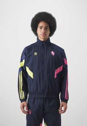 Giacca a vento navy con strisce geometriche gialle e rosa, con logo di un leone rosa, abbinata a pantaloni coordinati. Tessuto liscio e leggero.