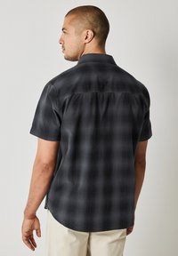 Kortärmad, mörk rutig skjorta med knappad krage. Slät yta och rundad nederkant med sidofickor för ökad rörlighet.