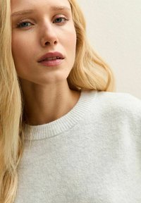 Pull en tricot gris clair avec un col rond côtelé, tissu texturé, porté par une personne aux longs cheveux blonds devant un fond uni.