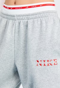 Svetlo sivi trenirka ima elastični pas z rdečo in belo logotipno trak. Na levem stegnu je bold rdeča vezenina "NIKE". Mehka tekstura tkanine.