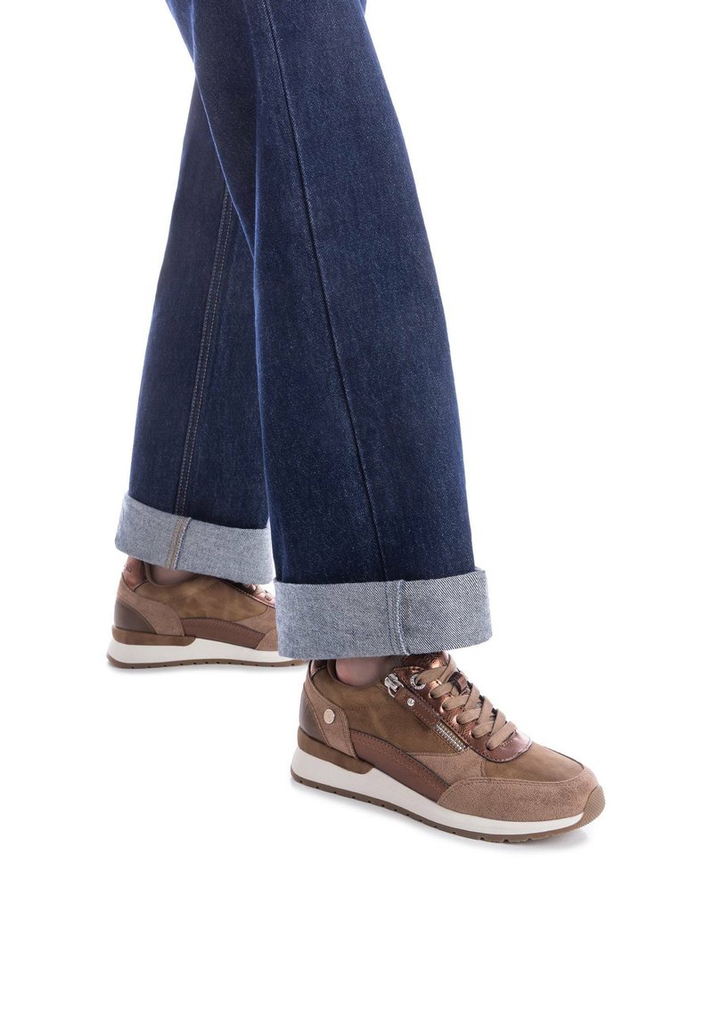 Zapatillas de ante marrón con acentos en bronce, cordones y cremalleras laterales. Combinadas con unos pantalones de mezclilla oscuros, remangados en los puños, que muestran un estilo casual.