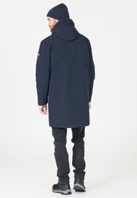 Parka azul marino con capucha, fabricada en material resistente al agua, con un corte más largo y detalles mínimos. Combinada con vaqueros oscuros y zapatos negros.