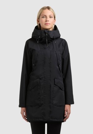 Mujer con abrigo parka negro con capucha, bolsillos delanteros, solapas con botones y cierre de cremallera, de pie frente a un fondo claro y liso.