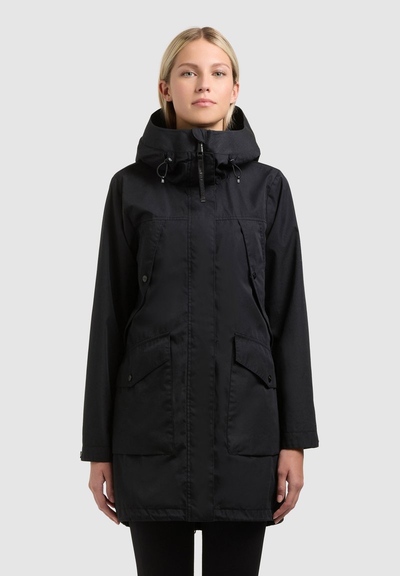 Femme portant un parka à capuche noir avec des poches avant, des rabats boutonnés et une fermeture éclair, debout devant un fond clair uni.