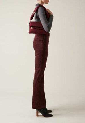 Borsa a tracolla bordeaux con finitura texture e una tracolla; indossata con un top grigio aderente e pantaloni bordeaux a vita alta, su uno sfondo neutro.