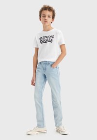 T-shirt bianca con logo nero Levi's, jeans azzurri chiari e sneakers beige con accenti rossi; vestibilità casual e trame di tessuto morbido.
