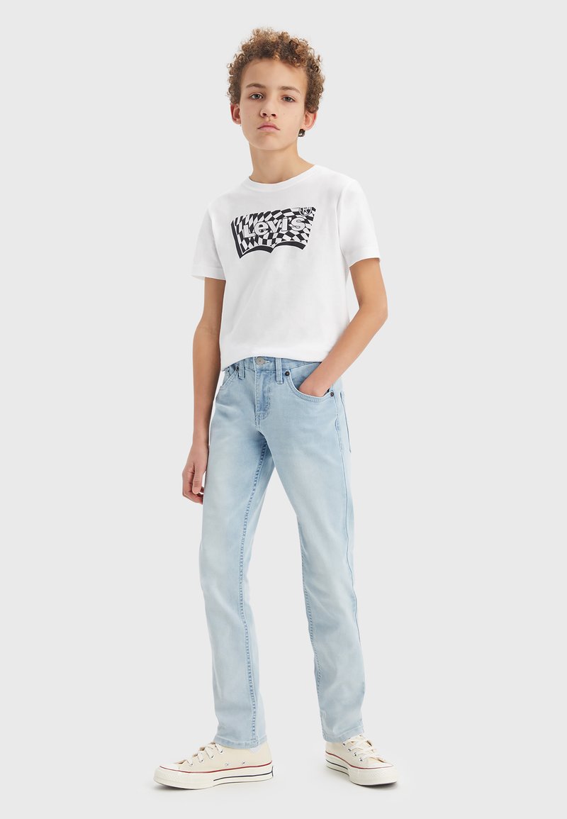 T-shirt bianca con logo nero Levi's, jeans azzurri chiari e sneakers beige con accenti rossi; vestibilità casual e trame di tessuto morbido.