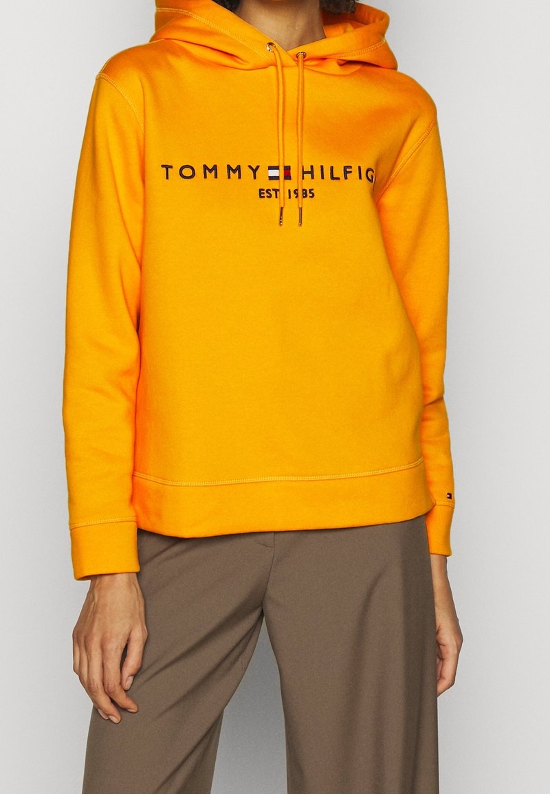 Personne portant un sweat à capuche Tommy Hilfiger orange vif avec logo et un pantalon beige, debout devant un fond clair uni.