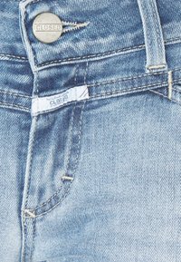 Primer plano de un bolsillo de unos jeans de mezclilla de color azul claro, mostrando un botón de metal y una pequeña etiqueta de tela blanca, ambos etiquetados como "CLOSED".