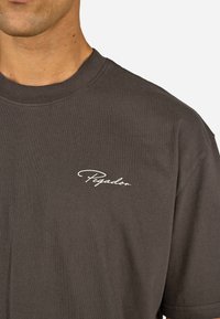 Camiseta de algodón gris oscuro con cuello redondo y mangas cortas, con un logo blanco de "Pegador" en el lado izquierdo del pecho.