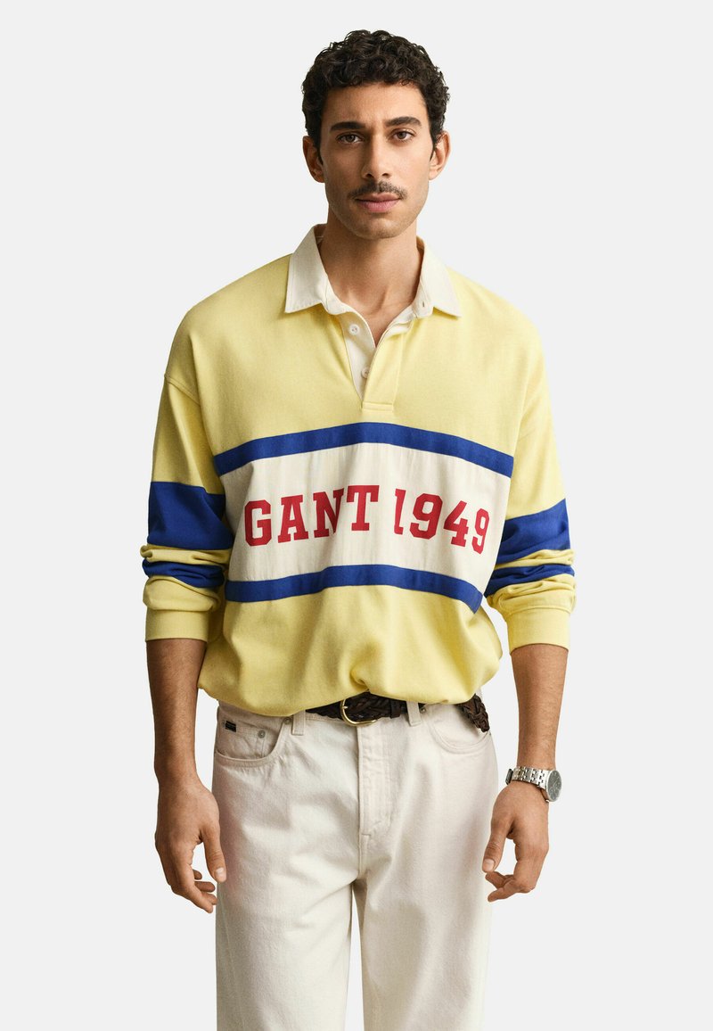 Uomo che indossa una polo a maniche lunghe gialla, blu e bianca con la scritta "GANT 1949", pantaloni bianchi, cintura intrecciata e orologio da polso argentato.