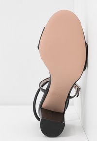 Sandale noire à talons hauts avec un dessus en cuir lisse, des brides fines et une semelle beige. Elle présente un bout pointu et une finition brillante.