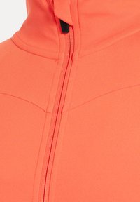 Koraalkleurig jack met ritssluiting, voorzien van een hoge kraag en een glad, rekbaar materiaal. Zichtbare naden en een contrasterende ritssluiting als detail.