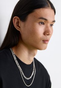 Sølvsmykker med forskellige kædestilarter hænger over en sort t-shirt, der fremhæver den glatte metaloverflade og lagdesign.