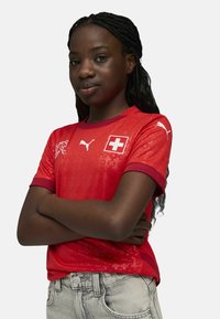 Punane lühikeste varrukatega jalgpallisärk Šveitsi lipu embleemiga, mustri tekstuuriga, ümmargune kaelus ja valge Puma logo õlgadel, kantud hallide teksadega.
