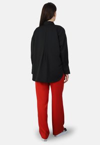 Camisa negra de manga larga con cuello alzado, costura en el centro de la espalda, combinada con pantalones rojos anchos y zapatos negros. Textura de tela suave.