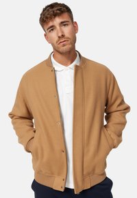 Béžová bomber bunda z vlněné směsi se zapínáním na zip, žebrovaným límcem a manžetami a bočními kapsami, nošená přes bílou polo tričko.