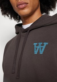 Brown hoodie with a front pocket, featuring a blue striped "W" logo embroidered at the chest, made of soft cotton material with a drawstring hood.