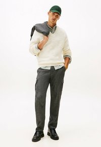 Joven con gorra verde, suéter blanco sobre camisa de rayas, pantalones cargo grises, zapatos negros, sosteniendo una chaqueta gris sobre el hombro, de pie sobre un fondo blanco.