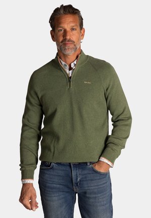 Man van middelbare leeftijd met zout-en-peper baard draagt een groen sweater met kwartrits over een bloemenhemd, gecombineerd met een blauwe spijkerbroek, staand met een hand in de zak.