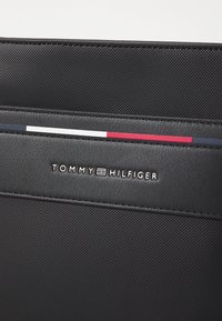 Mustapintainen lompakko, jossa on "TOMMY HILFIGER" -logo. Reunassa on kolmivärinen koristeviiva punaisena, valkoisena ja sinisenä.