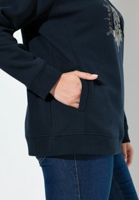 Sudadera azul marino con un diseño texturizado en la parte frontal, que cuenta con un bolsillo tipo canguro y puños acanalados. Combinada con jeans de mezclilla.