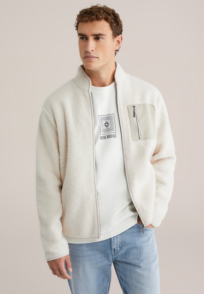 Chaqueta de forro polar en color crema con cuello alto, cierre de cremallera y un bolsillo gris claro en el pecho. Llevada sobre una sudadera blanca con gráfico.