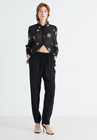 Chemise noire transparente à boutons avec broderies florales, associée à un pantalon noir taille haute. Talons transparents soulignés par un design simple.
