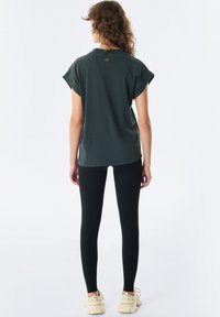 Ciemnozielony t-shirt z krótkim rękawem z podwiniętymi mankietami, luźny krój i detal z tyłu. Połączony z czarnymi legginsami i jasnymi sneakersami.