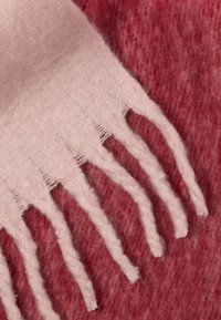 Cachecol de lã rosa suave com franjas, dobrado sobre um fundo de tecido de lã vermelho texturizado.