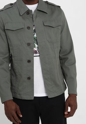 Mand iført olivengrøn knappet jakke med klaplommer på brystet over en hvid grafisk T-shirt og sorte bukser, der står mod en ensfarvet baggrund.
