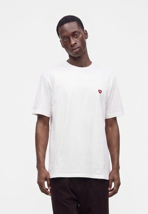 Jeune homme à la peau foncée portant un t-shirt blanc uni avec un petit emblème en forme de cœur rouge sur la poitrine, sur fond blanc.