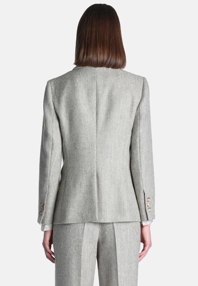 Personne aux cheveux bruns raides portant une veste de costume gris clair à motif chevrons et un pantalon assorti, vue de dos.