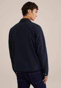 Marineblauwe lichtgewicht jas met een klassieke kraag, lange mouwen en knoopsommen, voorzien van een gladde, matte textuur. Gestyled met donkere denim.