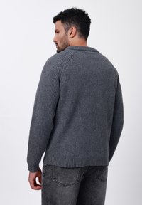 Grå ribbad stickad tr�öja med vikkrage, långa ärmar och avslappnad passform, tillsammans med mörkgrå jeans. Texturerad tygdetalj synlig.
