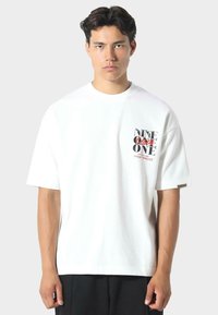 Mladý muž s krátkými tmavými vlasy nosí bílý oversize tričko s černým a červeným textem na hrudi a stojí na světle šedém pozadí.