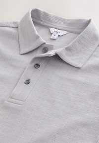Polo grigio chiaro con tessuto strutturato, dotata di abbottonatura e colletto strutturato. Etichetta del marchio visibile all'interno del colletto.