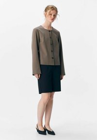 Veste marron à boutons, manches longues, en tissu doux ; associée à un short noir ajusté et des chaussures à talons noires. Design minimaliste.